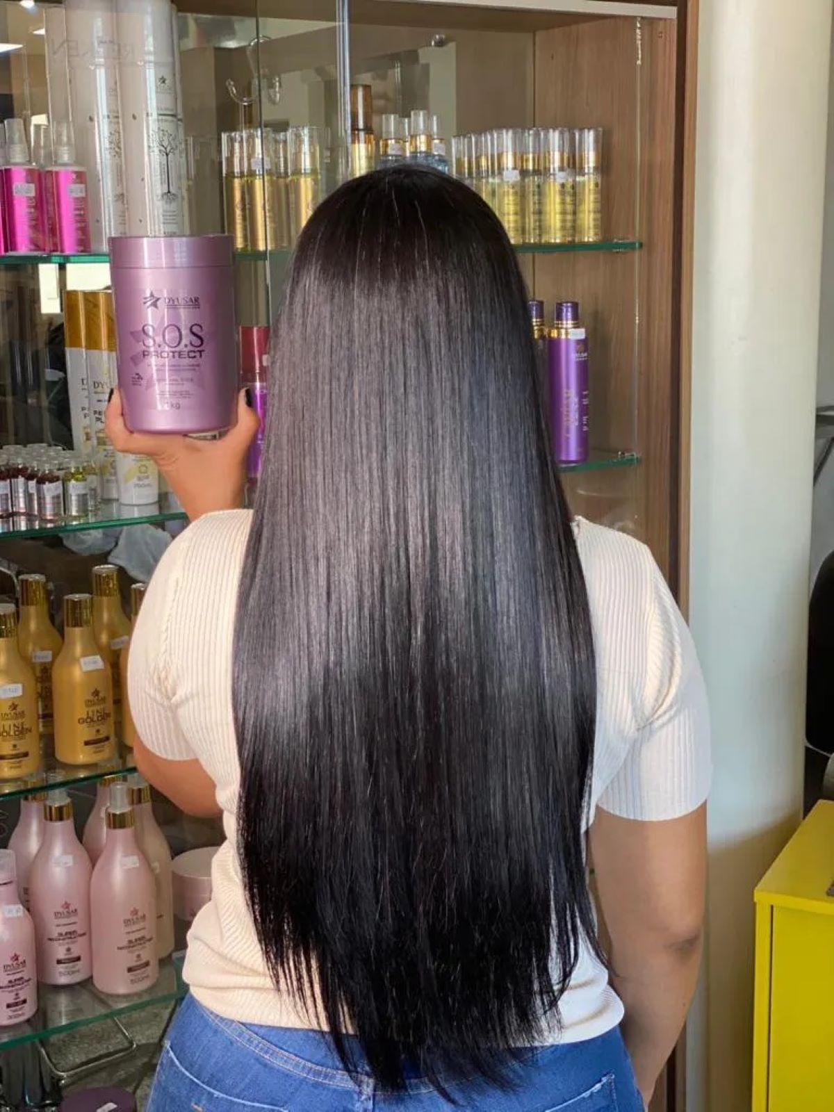 Cliente em um salão de beleza, vista de costas, mostrando o resultado de brilho intenso em seu cabelo longo e preto, enquanto segura o pote da Máscara Antioxidante S.O.S Protect.