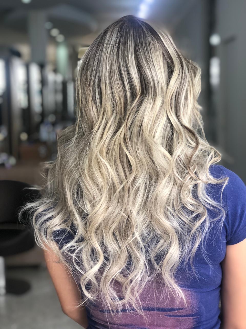 Mulher vista de costas em um salão, exibindo cabelo longo e ondulado, com luzes loiras saudáveis e brilhantes após o uso da Máscara Antioxidante S.O.S Protect.