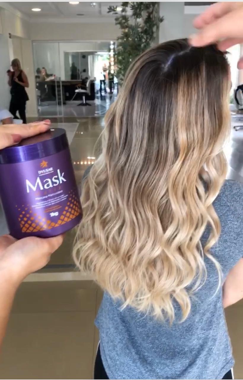 Profissional de salão segurando o pote de 1kg da Máscara Matizadora Dyusar ao lado do cabelo de uma cliente com longas ondas e luzes ombré.