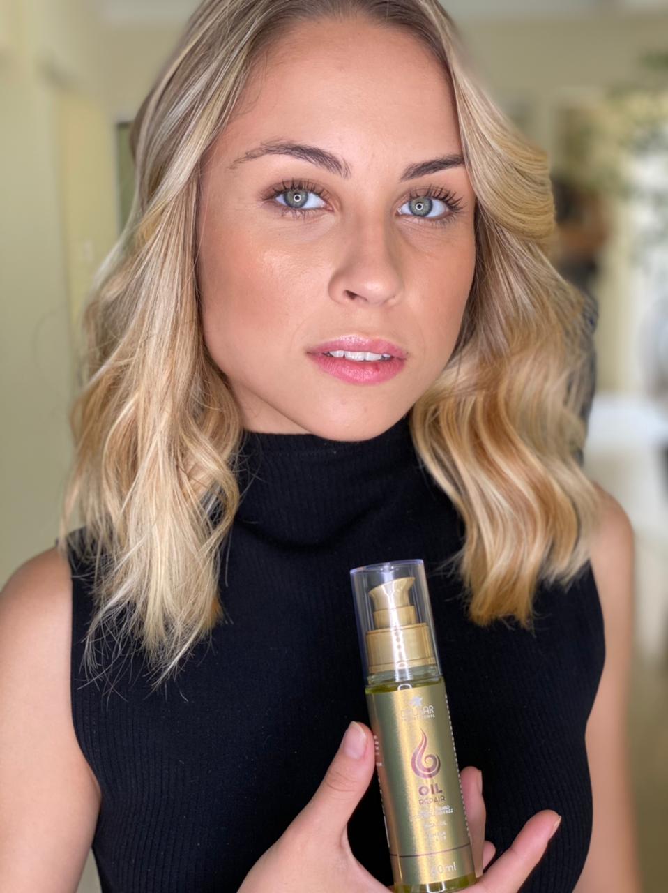 Modelo loira de olhos claros e cabelo ondulado, segurando o frasco do Óleo Reparador de Pontas Dyusar Oil Repair.