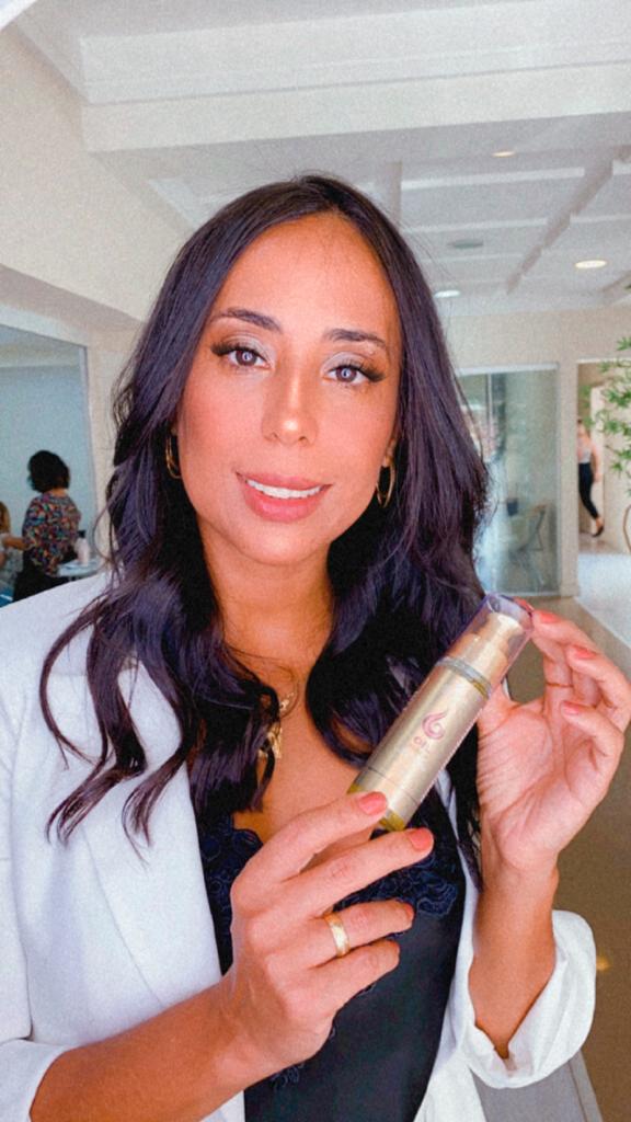 Mulher de blazer branco e cabelo escuro ondulado segura e apresenta o frasco do Óleo Reparador de Pontas Line Golden da Dyusar.