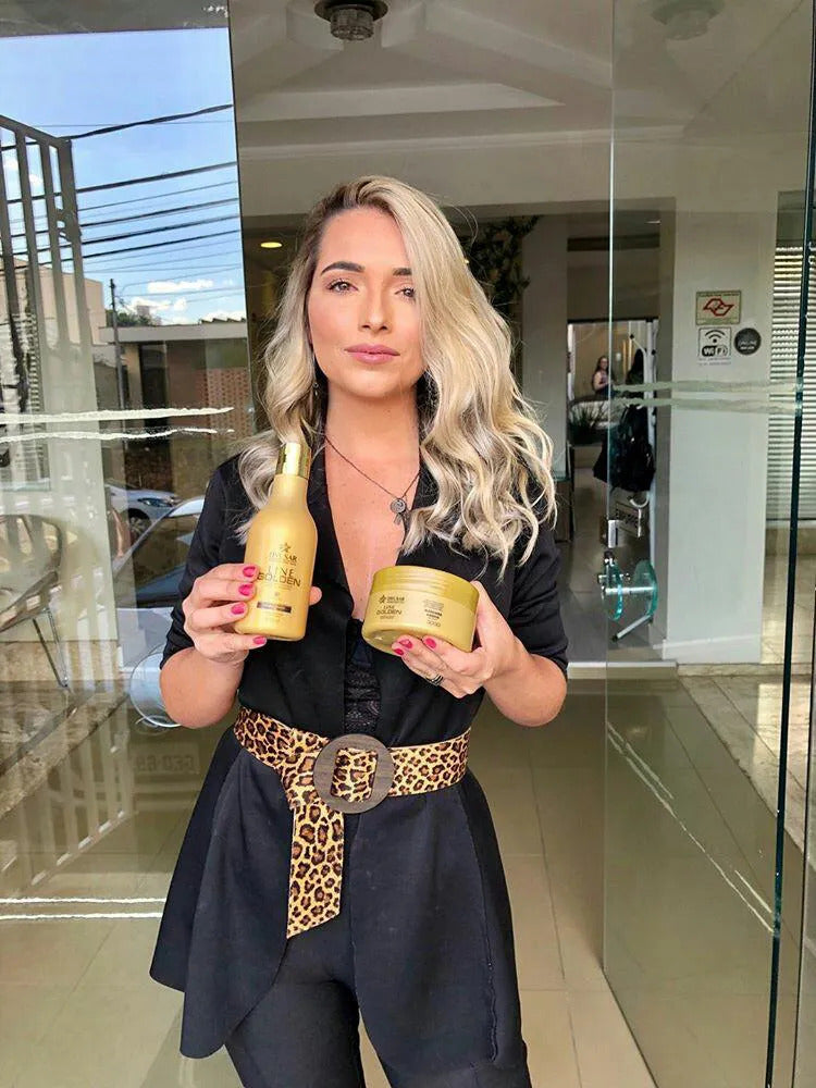 Mulher loira e estilosa com um cinto de oncinha, em pé na porta de um salão, mostrando o kit com o shampoo e a Máscara de Hidratação Dyusar Line Golden.
