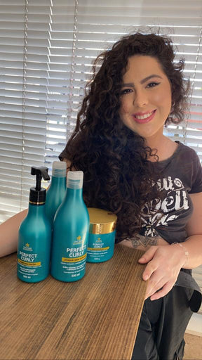 Mulher sorridente com longos cabelos cacheados, exibindo a linha completa de produtos Perfect Curly, incluindo o Shampoo para Cabelos Cacheados, sobre uma mesa de madeira.