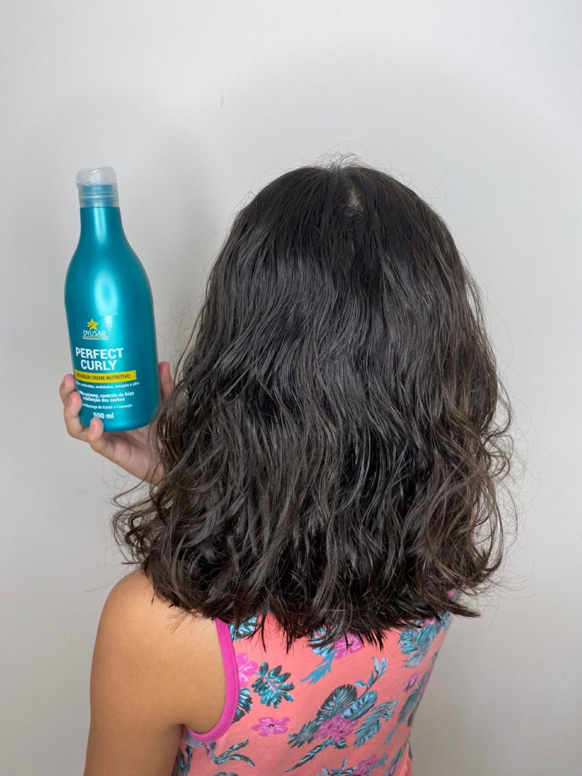 Criança de costas com cabelo ondulado e molhado, segurando o frasco do Ativador de Cachos da linha Perfect Curly, mostrando a aplicação do produto.

