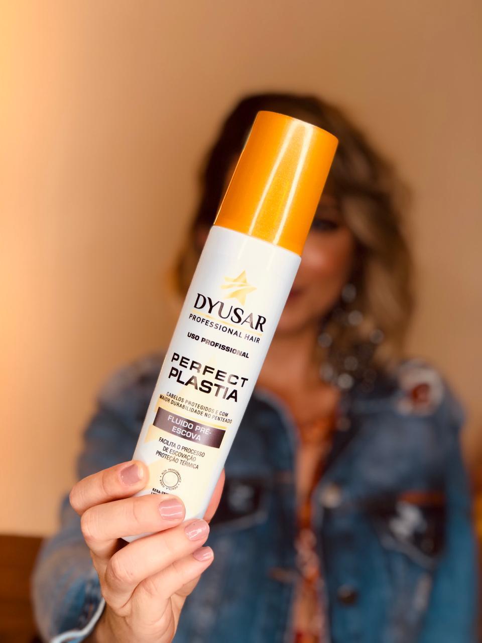 Mão segurando o frasco do Protetor Térmico e Fluido Pré-Escova Perfect Plastia em primeiro plano, com uma mulher desfocada ao fundo.