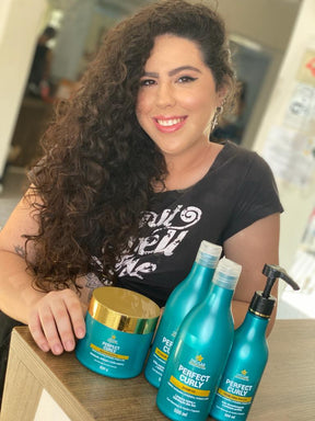 Cliente sorridente com cabelo cacheado longo e volumoso, apresentando o kit completo da linha Perfect Curly, que inclui o Shampoo para Cabelos Cacheados.