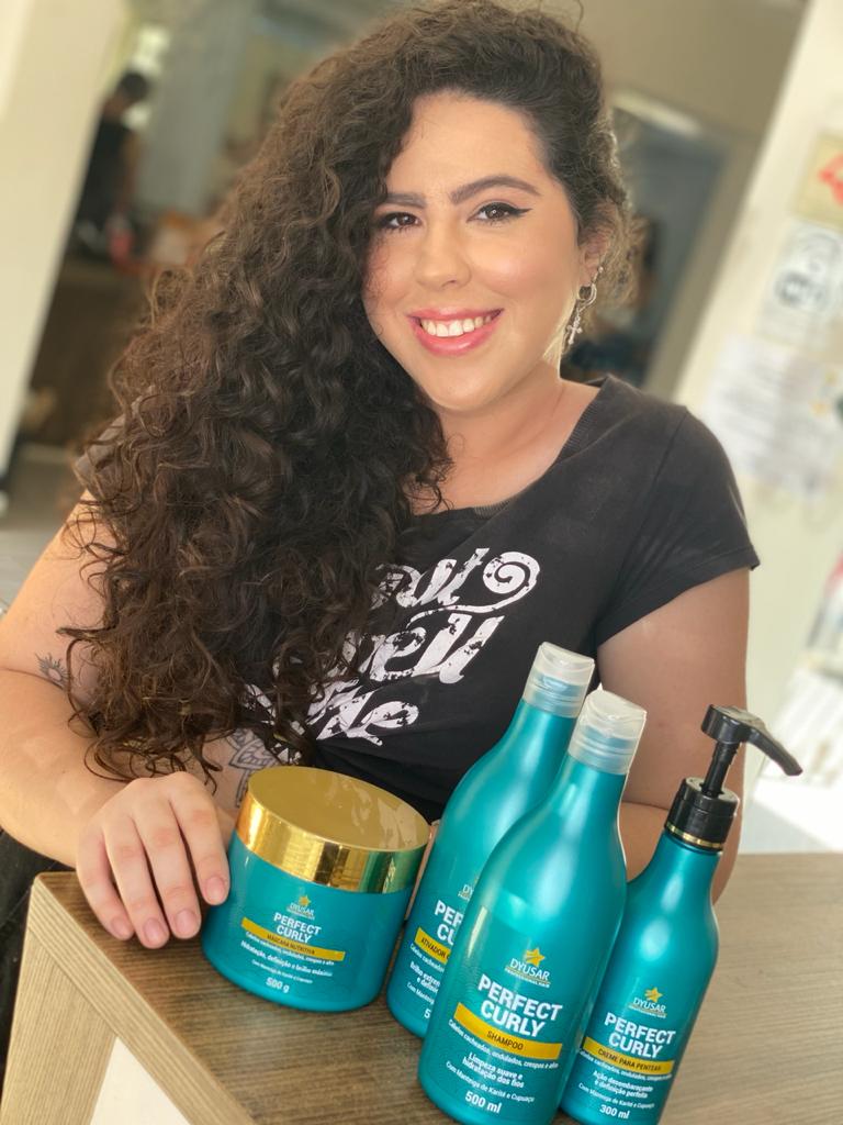 Cliente sorridente com cabelo cacheado longo e volumoso, apresentando o kit completo da linha Perfect Curly, que inclui o Shampoo para Cabelos Cacheados.