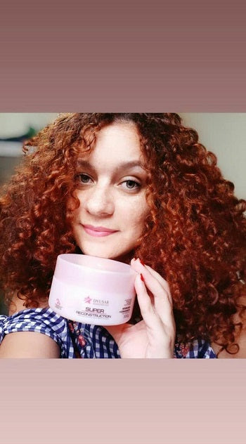 Modelo com cabelo cacheado ruivo segurando a Máscara de 300g do Kit Home Care Super Reconstruction.