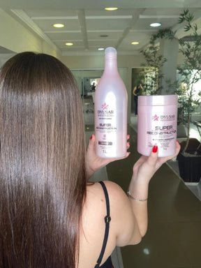 Mulher de costas em um salão exibindo cabelo liso e com brilho, enquanto segura o shampoo de 1L e a máscara de 1kg da linha Super Reconstruction, que também possui o Condicionador Reconstrutor.

