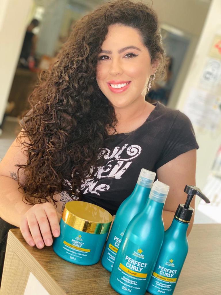 Cliente sorrindo com cabelo cacheado longo e volumoso, posando ao lado da linha completa Perfect Curly, que inclui o Creme de Pentear Para Cachos.