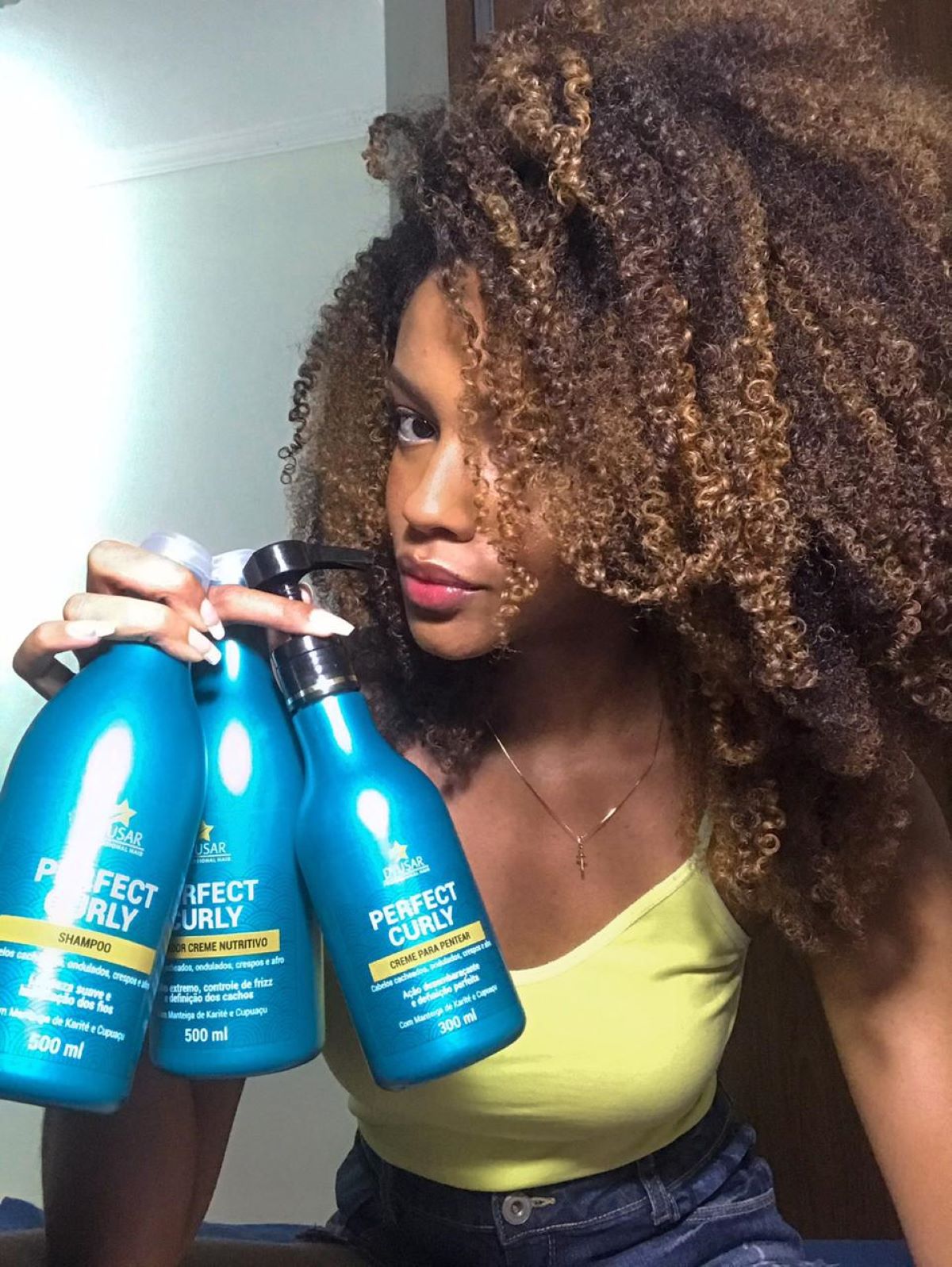 Modelo com cabelo crespo e luzes segurando a linha de produtos Perfect Curly, que inclui o Ativador de Cachos para definição e nutrição.

