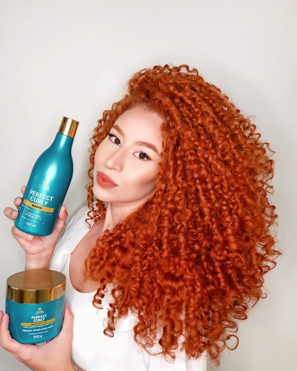 Modelo com cabelo cacheado, volumoso e ruivo, segurando o shampoo e a máscara da linha Perfect Curly, que prepara o cabelo para o uso do Ativador de Cachos.

