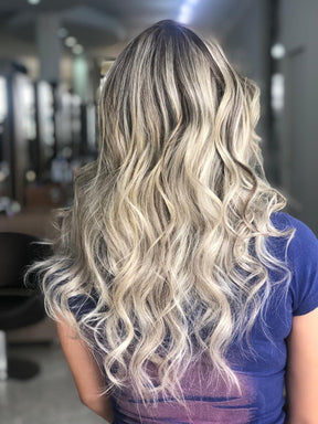 Mulher vista de costas em um salão, exibindo cabelo longo e ondulado, com luzes loiras saudáveis e brilhantes após o uso da Máscara Antioxidante S.O.S Protect.