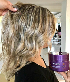 Mulher em um salão de beleza mostrando o resultado de seu cabelo loiro médio, ondulado e com luzes, enquanto segura o pote de 300g da Máscara Matizadora Dyusar.