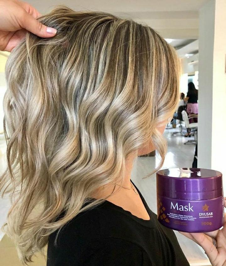 Mulher em um salão de beleza mostrando o resultado de seu cabelo loiro médio, ondulado e com luzes, enquanto segura o pote de 300g da Máscara Matizadora Dyusar.