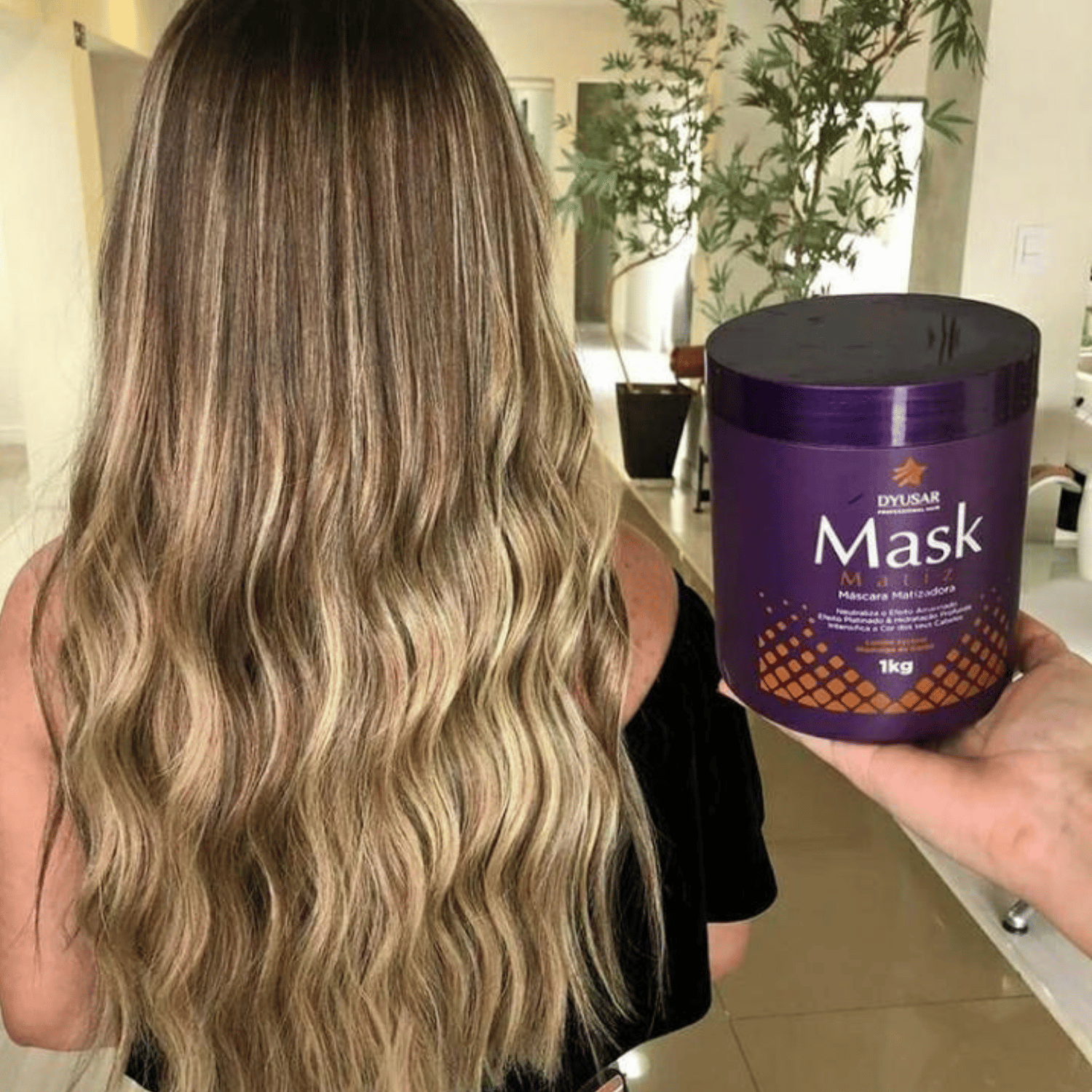 Cabelo longo e ondulado de uma mulher com mechas loiras, ao lado de uma mão segurando o pote de 1kg da Máscara Matizadora Dyusar.