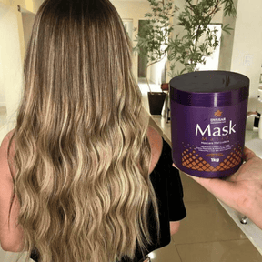 Cabelo longo e ondulado de uma mulher com mechas loiras, ao lado de uma mão segurando o pote de 1kg da Máscara Matizadora Dyusar.