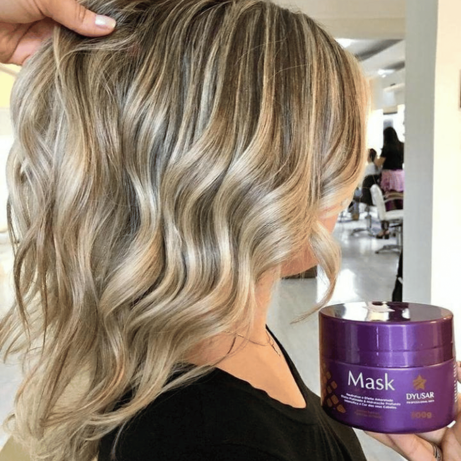 Cabelo loiro e ondulado com mechas sendo segurado por uma mão, e pote da Máscara Matizadora Dyusar à frente.