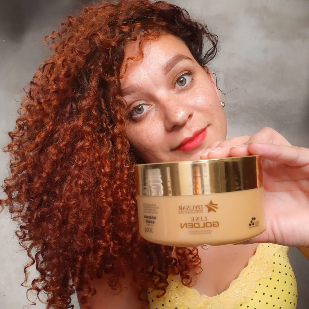 Modelo com cabelo cacheado ruivo mostrando a Máscara Line Golden (300g), a etapa de Hidratação do Kit Cronograma Capilar.