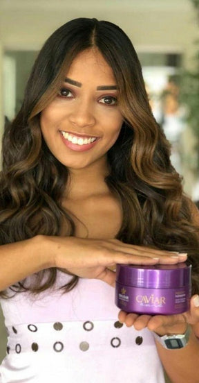 Modelo sorrindo com cabelo longo, ondulado e com luzes (morena iluminada), segurando o pote da Máscara de Nutrição com Caviar da Dyusar.