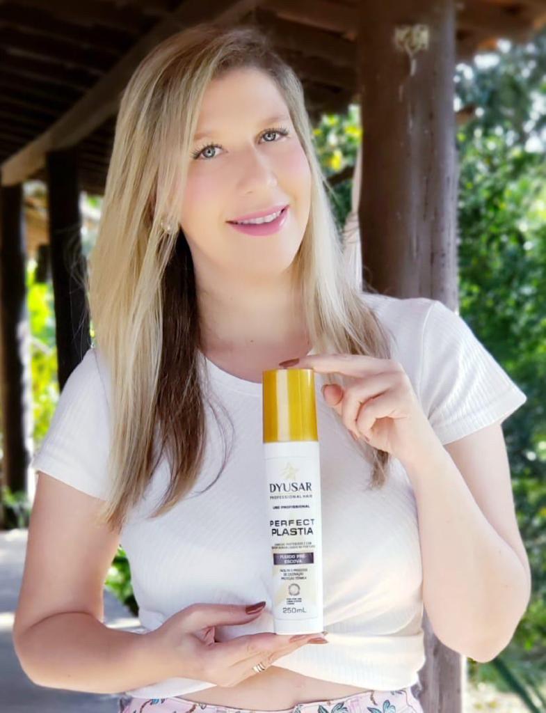 Mulher loira sorrindo em um ambiente externo, segurando e mostrando o frasco do Protetor Térmico e Fluido Pré-Escova da linha Perfect Plastia.