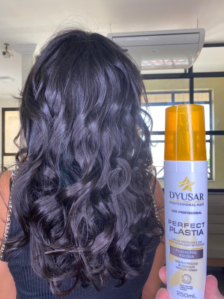 Resultado de cabelo escuro, ondulado e com muito brilho em um salão, com o frasco do Protetor Térmico e Fluido Pré-Escova da Dyusar ao lado.