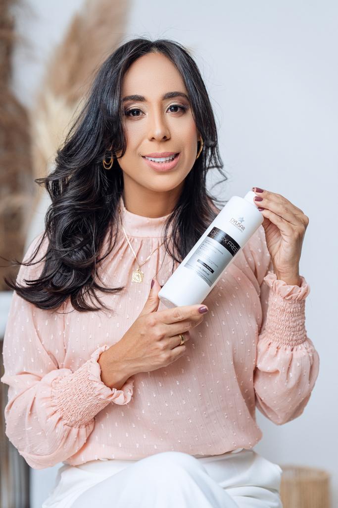 Mulher sorridente com cabelo escuro e ondulado, vestindo uma blusa rosa, segura e apresenta o frasco do Shampoo Detox da Dyusar.