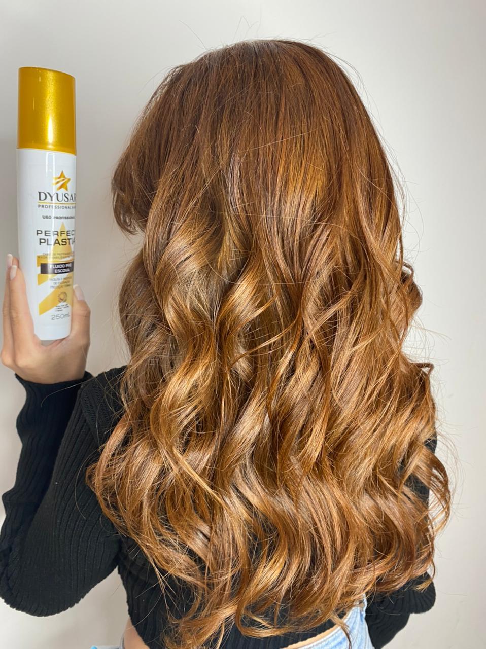Mulher de costas com cabelo longo, ruivo e ondulado, mostrando o resultado de brilho e definição enquanto segura o frasco do Protetor Térmico e Fluido Pré-Escova Dyusar.